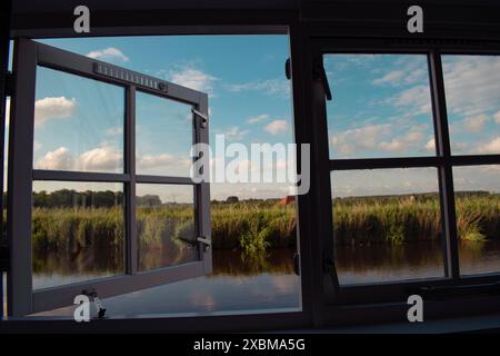 Une fenêtre ouverte donnant sur un paysage tranquille avec un canal, des prairies et un ciel nuageux, Ilpendam, pays-Bas Banque D'Images