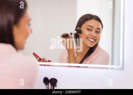 Femme biraciale heureuse appliquant le maquillage tout en souriant à son reflet dans le miroir Banque D'Images