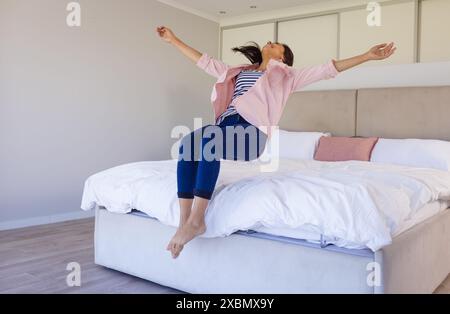 Femme biraciale heureuse sautant sur le lit avec les bras tendus, regardant joyeuse Banque D'Images