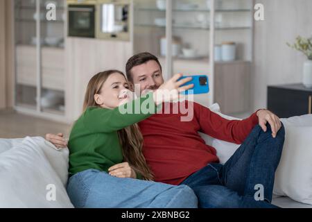 Couple joyeux assis sur le canapé, regardant dans la caméra de smartphone, faisant selfie pour l'envoi aux parents Banque D'Images