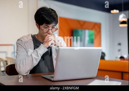 Un jeune homme asiatique réfléchi portant des lunettes et des vêtements décontractés travaille sur son ordinateur portable, regardant l'écran avec un visage sérieux, pensez Banque D'Images