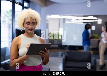 Jeune femme biraciale utilisant la tablette au bureau comme les collègues discutent en arrière-plan, copiez l'espace Banque D'Images