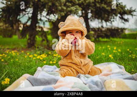 Mignon bébé fille en costume en peluche avec pomme rouge dans les mains assis sur une couverture dans l'herbe verte Banque D'Images