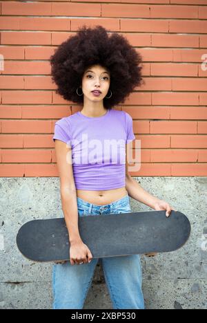 Confiante jeune femme afro-américaine avec coiffure afro dans des vêtements décontractés debout avec skateboard contre le mur de briques et regardant la caméra Banque D'Images