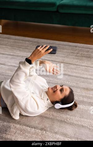 Femme souriante appel vidéo dans les vêtements de loisirs à la maison, portant des écouteurs et en utilisant un smartphone, copiez l'espace Banque D'Images