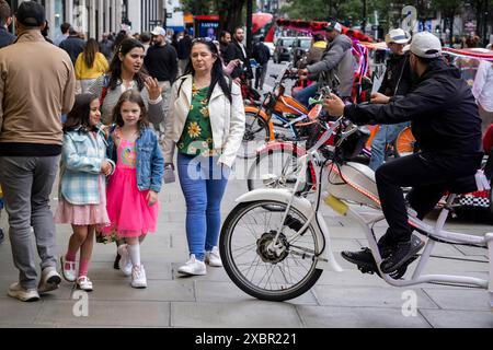 Acheteurs et visiteurs sur Oxford Street devant le magasin Selfridegs alors que les cyclistes-taxis attendent en ligne sur le trottoir pour les tarifs le 9 juin 2024 à Londres, Royaume-Uni. Oxford Street est un grand centre commercial dans le West End de la capitale et est la rue commerçante la plus fréquentée d'Europe avec environ un demi-million de visiteurs quotidiens dans ses quelque 300 magasins, dont la majorité sont des magasins de mode et de vêtements de grande rue. Banque D'Images