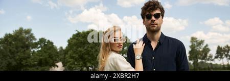 Un homme et une femme élégants, habillés avec élégance, se tiennent ensemble dans un champ vert éclatant, tous deux portant des lunettes de soleil. Banque D'Images