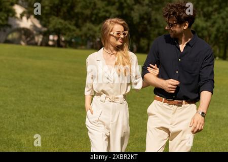 Un beau jeune couple en vêtements élégants marche ensemble sur un champ vert, incarnant un style de vie luxueux et ancien. Banque D'Images