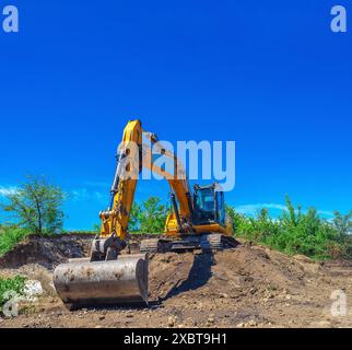 Excavatrice sur chenilles en pente contre ciel bleu. Défrichement, creusement de fondations, travaux de construction Banque D'Images