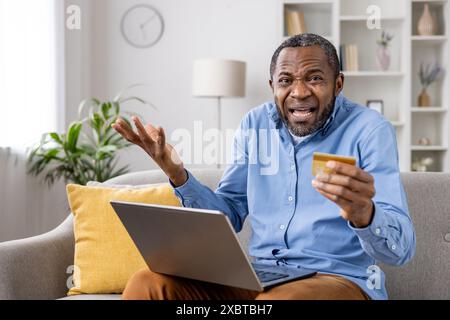 Un homme confus tenant une carte de crédit tout en faisant des achats en ligne, manifestant des inquiétudes au sujet de la fraude sur Internet et des escroqueries en ligne. Banque D'Images