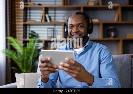 Homme heureux portant des écouteurs à l'aide d'une tablette tout en étant assis dans un environnement de salon confortable. Le cadre comprend des étagères et de la verdure, suggérant une atmosphère détendue et confortable. Banque D'Images