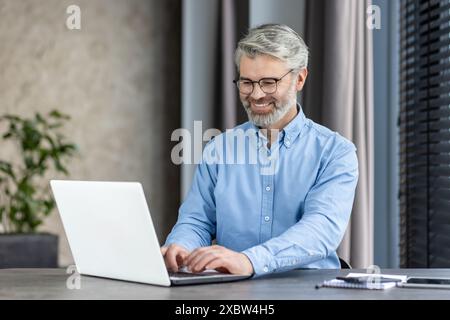 Homme âgé avec des lunettes et une barbe souriant tout en utilisant un ordinateur portable dans un environnement de bureau moderne Banque D'Images