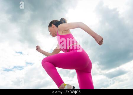 Vue en angle bas d'une femme en train de courir, portant des vêtements de sport roses contre un ciel spectaculaire. Concept fitness. Banque D'Images