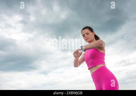 Femme sportive vérifiant sa montre de tracker de fitness, portant des vêtements de sport roses contre un ciel dramatique. Banque D'Images