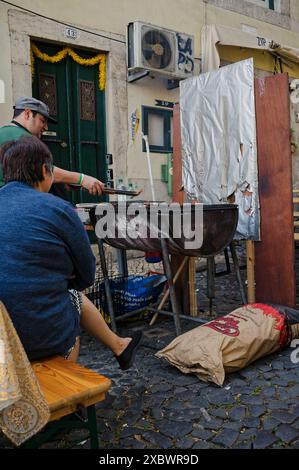 Alfama pendant les festivités populaires des saints. Les habitants préparent un grill au charbon de bois dans une rue pavée avec des décorations festives suspendues Banque D'Images