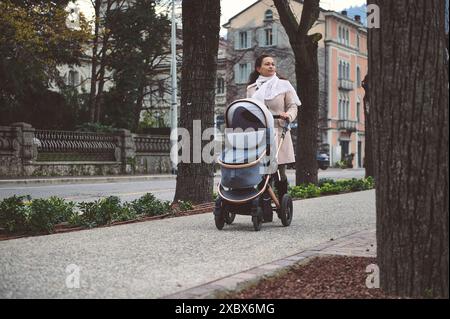 Une jeune mère se promène tranquillement avec une poussette de bébé sur un trottoir de la ville en automne, profitant de l'environnement urbain serein. Banque D'Images
