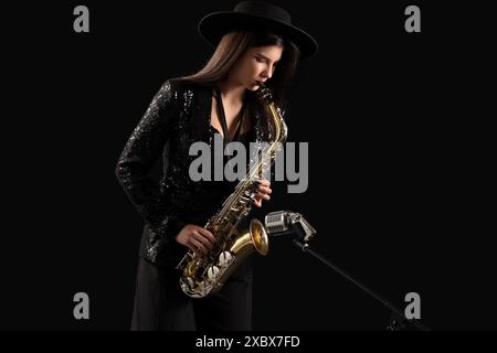 Belle jeune femme avec microphone jouant du saxophone sur fond sombre Banque D'Images