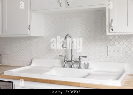 Un détail de robinet de cuisine avec un grand évier de ferme blanc avec doubles égouttoirs, carreaux de dosseret arabesque et comptoir de bloc de boucher naturel. Banque D'Images