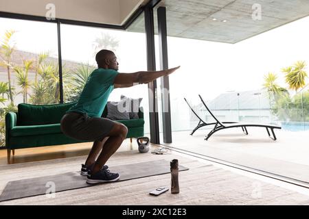 Ajuster afro-américain masculin squatting sur tapis de yoga dans le salon moderne, l'exercice Banque D'Images