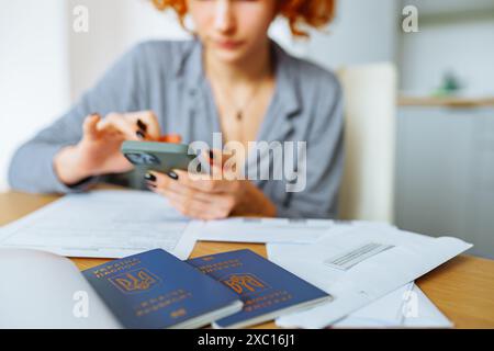 Jeune femme, étudiante, avec les cheveux roux bouclés, remplit des documents assis à table à la maison, au premier plan est citoyen de passeport de l'Ukraine Banque D'Images