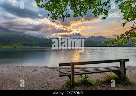 Un banc en bois se trouve sur la rive d'Almsee, surplombant un lac serein avec en toile de fond des montagnes majestueuses. Le soleil couchant projette une lueur chaude à travers Banque D'Images