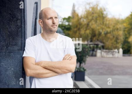 Homme en t-shirt blanc debout avec les bras croisés appuyés contre le mur Banque D'Images