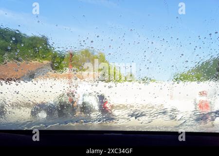 Vue de la rue à travers un pare-brise de voiture recouvert de gouttes d'eau. Photo de l'intérieur d'un véhicule dans un lave-auto. Banque D'Images