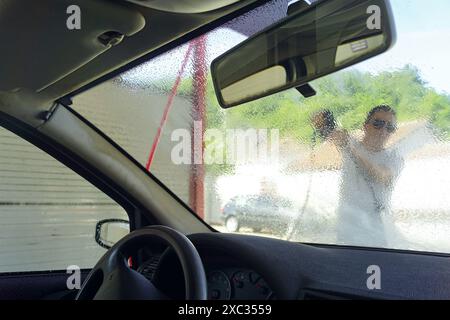 Un homme lave manuellement une voiture dans un lave-auto à ciel ouvert - photo prise de l'intérieur de la voiture depuis le siège passager. Banque D'Images