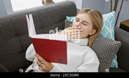Une femme caucasienne fatiguée bâillant tout en lisant un livre rouge à l'intérieur, assise confortablement dans un salon moderne. Banque D'Images