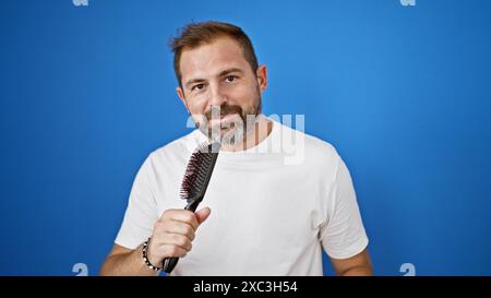 Bel homme mature avec les cheveux gris souriant tenant une brosse à cheveux sur un fond bleu en plein air. Banque D'Images