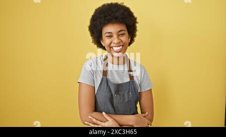 Femme noire souriante avec les cheveux bouclés et les bras croisés portant un tablier sur un fond jaune. Banque D'Images