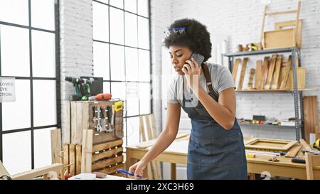 Femme afro-américaine charpentier multitâche avec téléphone dans un atelier bien éclairé Banque D'Images