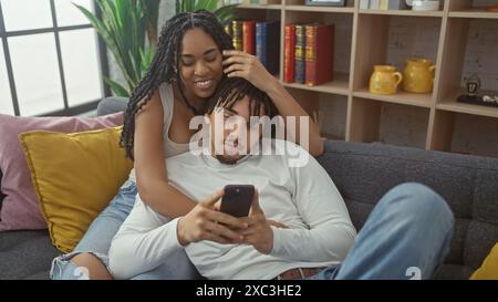 Une femme caresse les cheveux de son partenaire pendant que l'homme utilise un smartphone, les deux se prélassant confortablement dans un cadre de salon confortable. Banque D'Images