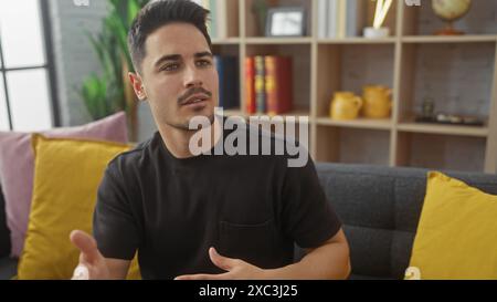 Un beau jeune homme hispanique avec une barbe dans une chemise noire parlant et faisant des gestes dans un salon moderne Banque D'Images