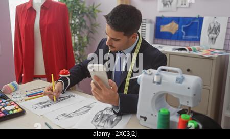 Un jeune homme hispanique avec une barbe dessine des dessins de mode dans un magasin de tailleur tout en vérifiant son téléphone. Banque D'Images