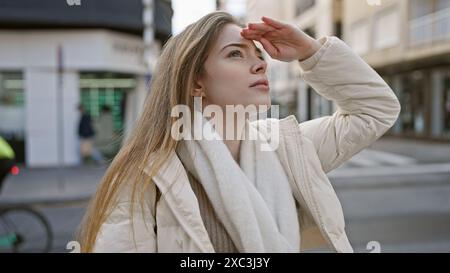 Une jeune femme blonde dans un cadre urbain protège ses yeux du soleil tout en se tenant debout sur une route. Banque D'Images