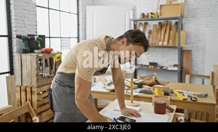 Jeune homme hispanique à l'artisanat de barbe dans un atelier de menuiserie ensoleillé, dégageant une ambiance artisanale qualifiée. Banque D'Images