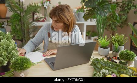 Femme concentrée d'âge moyen prenant des notes dans une boutique de fleuriste luxuriante entourée de plantes et d'un ordinateur portable. Banque D'Images