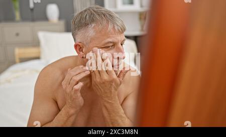 Un homme pensif d'âge moyen touchant son visage alors qu'il était assis dans son lit, représente la routine du matin ou les soins de la peau dans un cadre de chambre. Banque D'Images