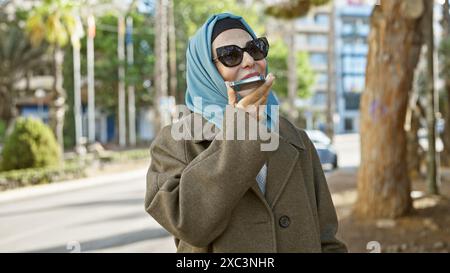 Femme musulmane en hijab utilisant un smartphone dans un parc urbain, illustrant un style de vie moderne et connecté. Banque D'Images