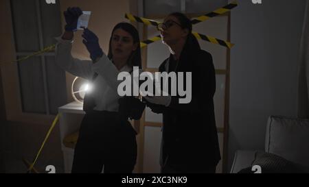 Deux enquêteurs femmes analysent les preuves à l'intérieur sur une scène de crime faiblement éclairée avec un ruban de mise en garde visible. Banque D'Images