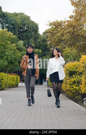 Deux femmes latines marchant dans un parc, elles ont quitté l'université et rentrent chez elles, image verticale, image verticale, elles se divertissent en regardant th Banque D'Images