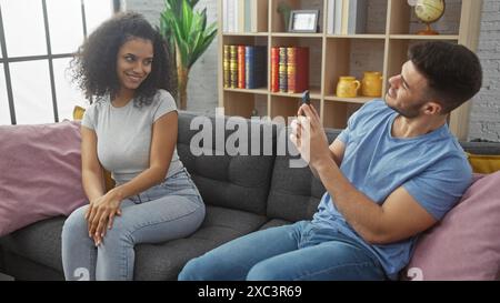 Un homme utilise un téléphone portable pour prendre une photo d'une femme souriante assise sur un canapé gris dans un salon confortable. Banque D'Images