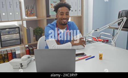 Beau jeune homme afro-américain avec une barbe et un stéthoscope assis dans un bureau de clinique souriant chaleureusement avec du matériel médical et des documents autour Banque D'Images
