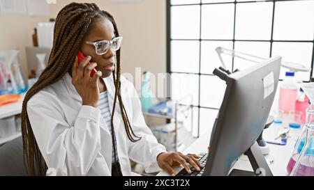 Femme afro-américaine en blouse de laboratoire à l'aide de l'ordinateur et du téléphone en laboratoire Banque D'Images