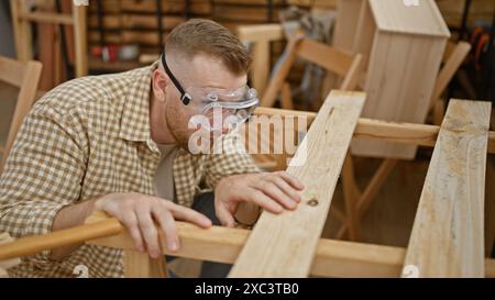 Un jeune homme concentré avec une barbe et des lunettes de sécurité sabre le bois dans un atelier bien éclairé Banque D'Images