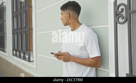 Un jeune homme adulte en tenue décontractée s’appuie contre un bâtiment urbain tout en utilisant son smartphone, dépeignant la vie quotidienne de la ville. Banque D'Images