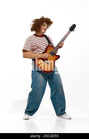 Jeune homme, musicien aux cheveux bouclés joue de la guitare électrique, entièrement immergé dans le rythme isolé sur fond blanc Banque D'Images