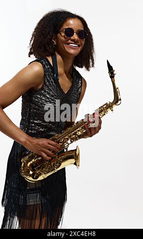 Élégance jazz. Portrait de belle jeune femme en robe noire et lunettes de soleil, posant avec un saxophone doré isolé sur fond blanc Banque D'Images