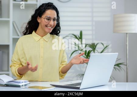Jeune femme hispanique souriante assise dans un bureau lumineux moderne, étudiant et travaillant sur un ordinateur portable, parlant sur un appel vidéo, faisant des gestes avec les mains. Banque D'Images
